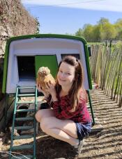 A happy customer holding her chicken on front of her green chicken coop