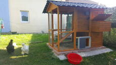 Een groot houten kippenhok met daaraan een grijze Autodoor in een tuin