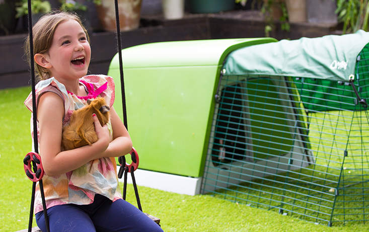 a girl sitting on a swing holding a chicken with a eglu go in the background